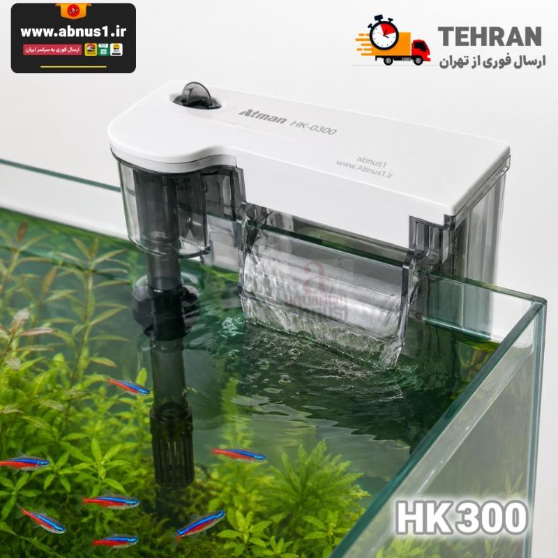 فیلتر آویزان برند “Atman” مدل HK-0300 است. در نمای روی جعبه، دستگاه با بدنه‌ای شفاف و مستحکم دیده می‌شود که بر روی لبه آکواریوم سوار شده است. در این مدل، فضای محفظه فیلتراسیون نسبت به مدل‌های پایه بزرگ‌تر شده که نویدبخش حجم مدیای بیشتر است. ویژگی‌های کلیدی مشهود در عکس شامل لوله مکش مجهز به چربی‌گیر سطح (Skimmer)، پیچ تنظیم شدت جریان آب در قسمت بالایی و خروجی آبشاری پهن است. همچنین روی جعبه به قابلیت تنظیم گیره نگهدارنده متناسب با ضخامت شیشه‌های مختلف اشاره شده است که نصب آن را بر روی هر تانکی آسان می‌کند.